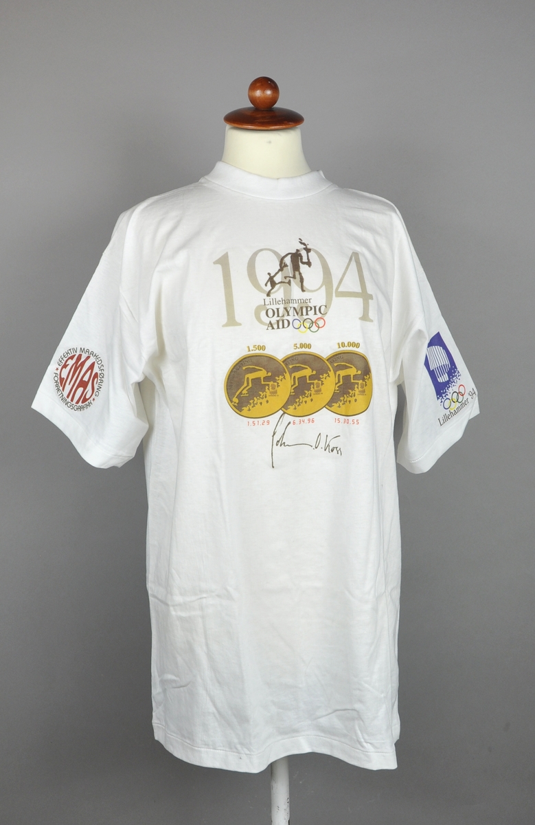 T-skjorte med logo for Lillehammer Olympic Aid foran teksten 1994. Det er også avbildet de tre gullmedaljene med angivelse av øvelse og tid, som Johan Olav Koss vant i 1994. På venstre erme er emblemet for Lillehammer '94, og på høyre erme logo for en samarbeidspartner.