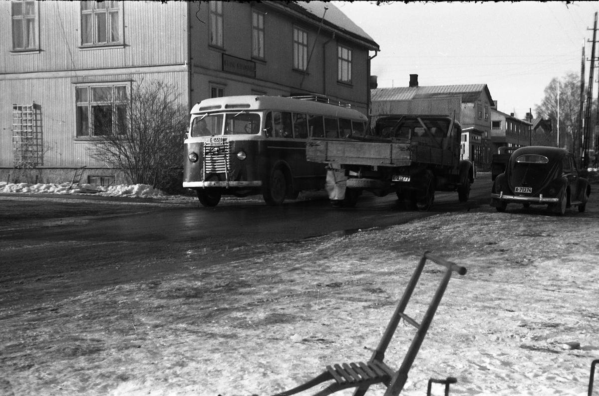 Bilde tatt oppover Lenagata mot jernbaneovergangen, gjennom vinduet fra Lena Foto & Radio i "Raubua" mars 1958. Grand til venstre i bildet. Bussen med kjennetegn E-15519, en Diamond T årsmodell 1947-48 med Sørholte karossseri, var en av bussene som i 1951 ble med inn i selskapet Østre Toten Bilruter (seinere endret til Bilselskap i stedet for Bilruter) som hadde hovedkontor på Skreia. I 1965 ble selskapet fusjonert inn i det nystartede Vestoppland Bilselskap. Bilen til høyre i bildet er en VW med oval bakrute, dvs. årsmodell 1953-57.