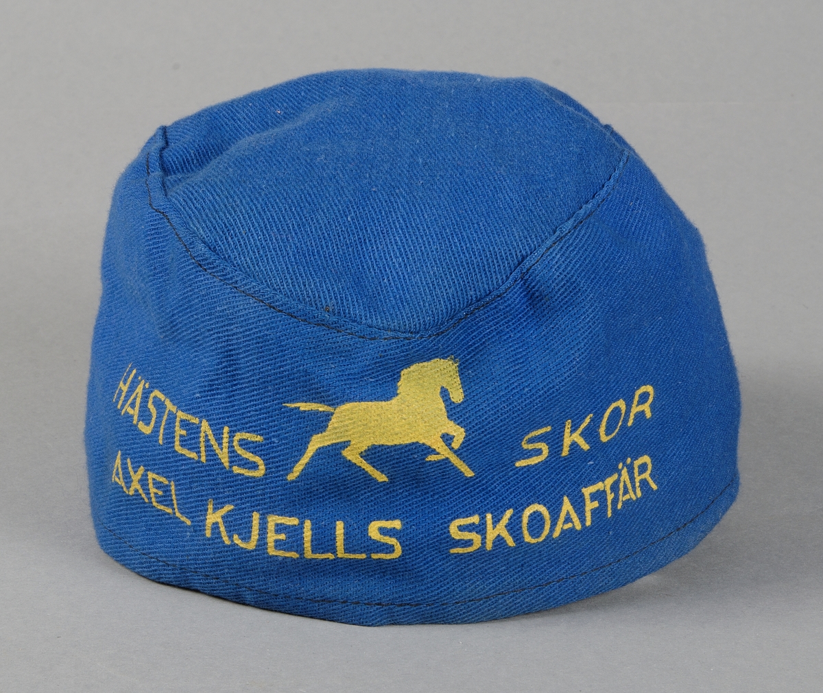 Båtmössa av blått bomullstyg med gul reklamtext: HÄSTENS SKOR, AXEL KJELLS SKOAFFÄR.
