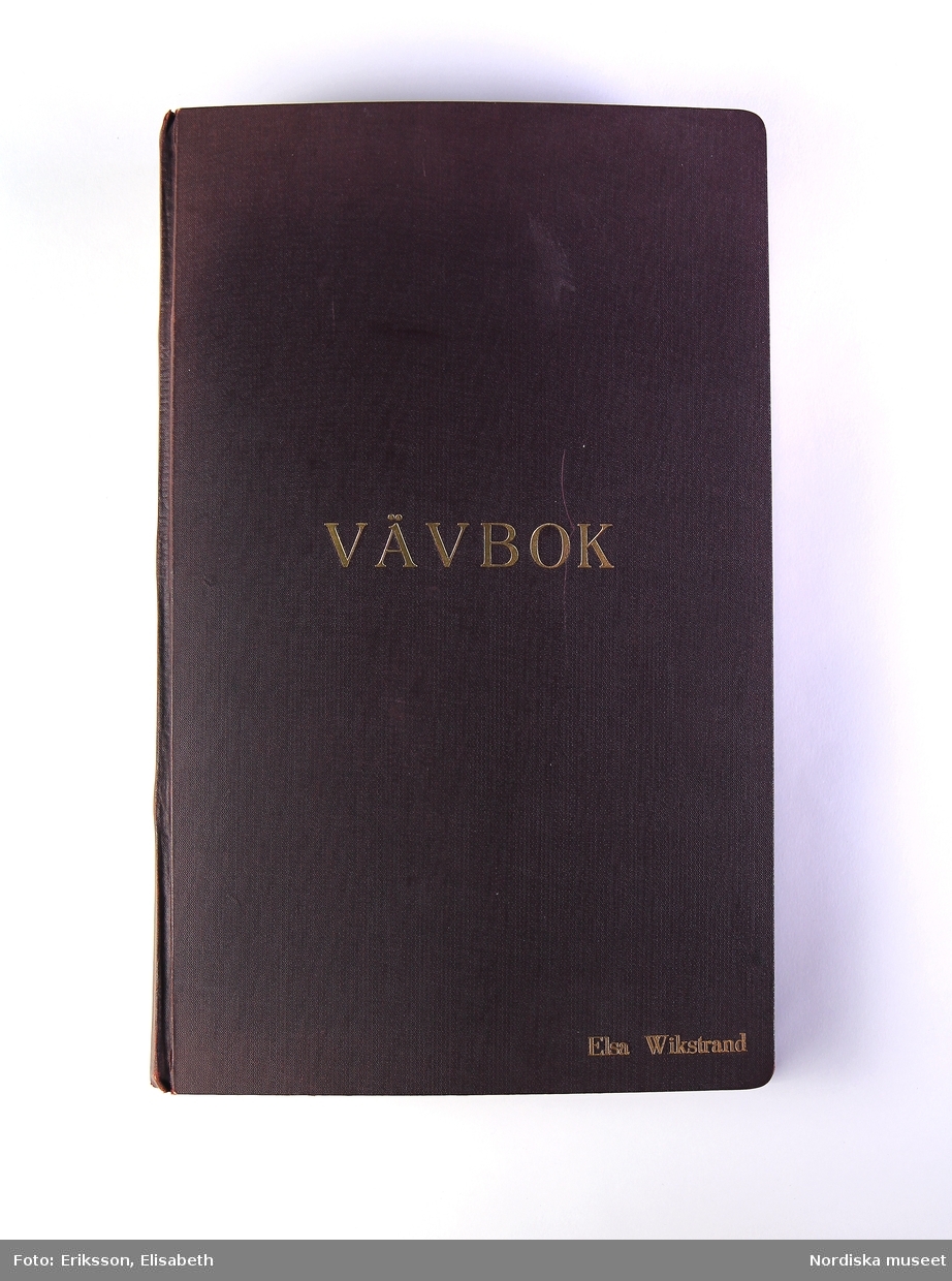 Vävbok. Svart pärm med tryck i guldfärg på framsidan "VÄVBOK" / "ELSA WIKSTRAND". Boken är handskriven med text och teckningar av vävstolar och vävnotor. Inklistrade tygprover.
