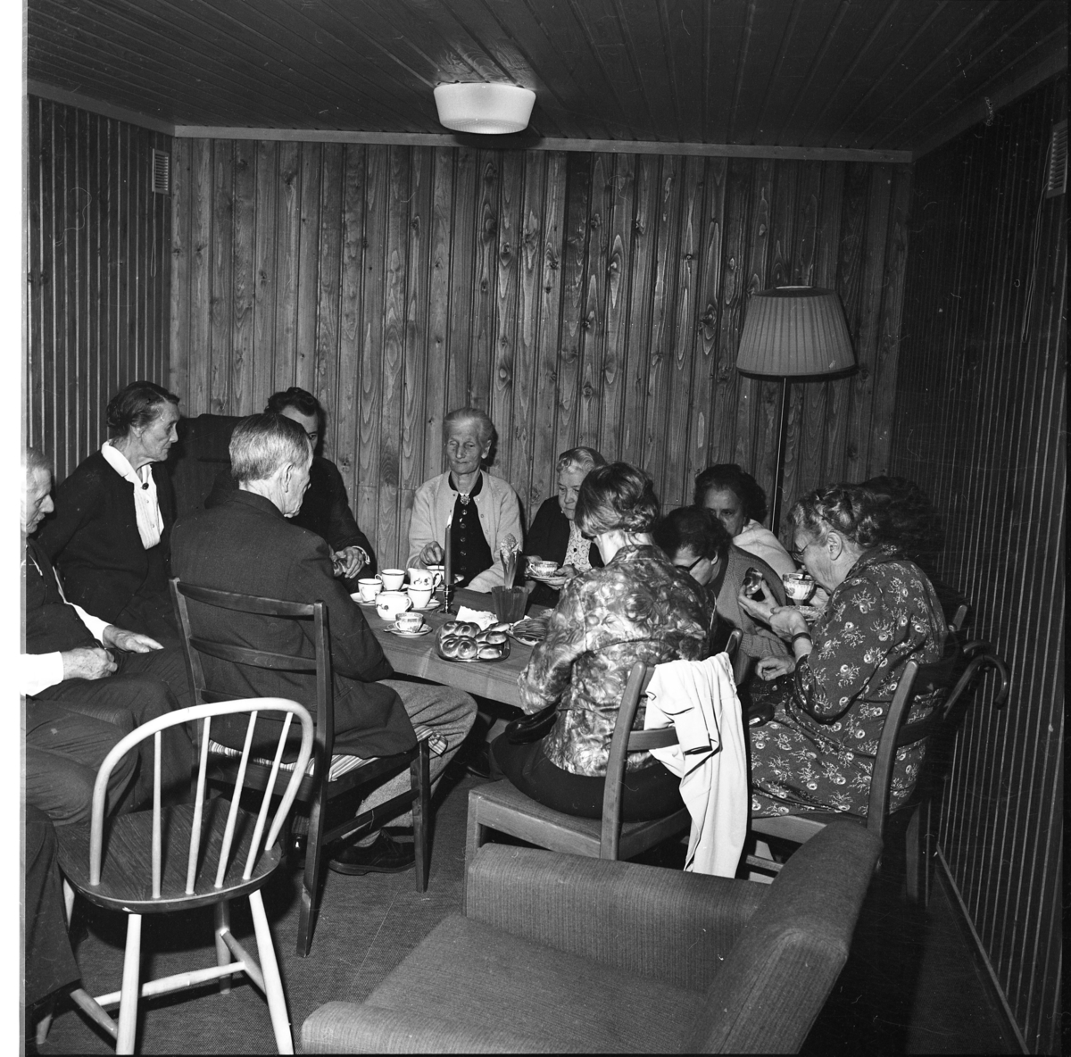 Äldre damer och herrar sitter omkring ett rektangulärt bord med duk, tända ljus samt kaffekoppar och kakfat. De fikar.