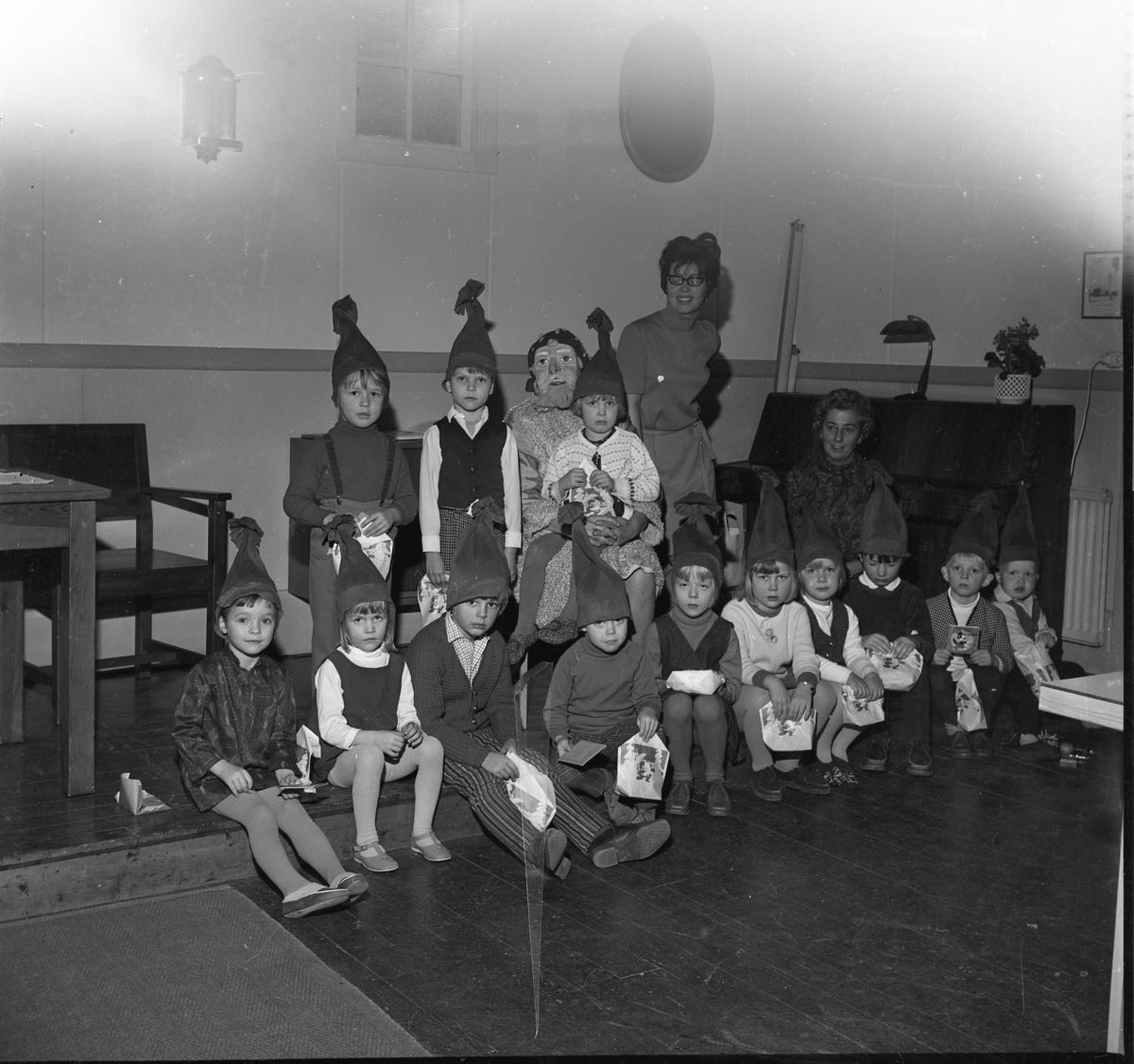 Julfest i Skärstad kyrka. Små flickor och pojkar sittande och stående med tomteluvor på huvudet. En flicka sitter i knät på en tomte. I bakgrunden står två kvinnlig lärare.