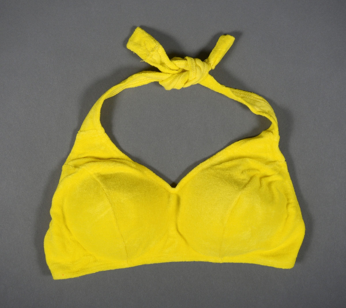 En gul bikinioverdel med halterneck (stroppene knytes i nakken). Den er laget av et flossete gult bomullsstoff. Cupen er polstret med et hvitt kunststoff. Midt bak er det en hekte av hvit plast som kan åpnes/lukkes. Bikinioverdelen er i størrelse 44/46.