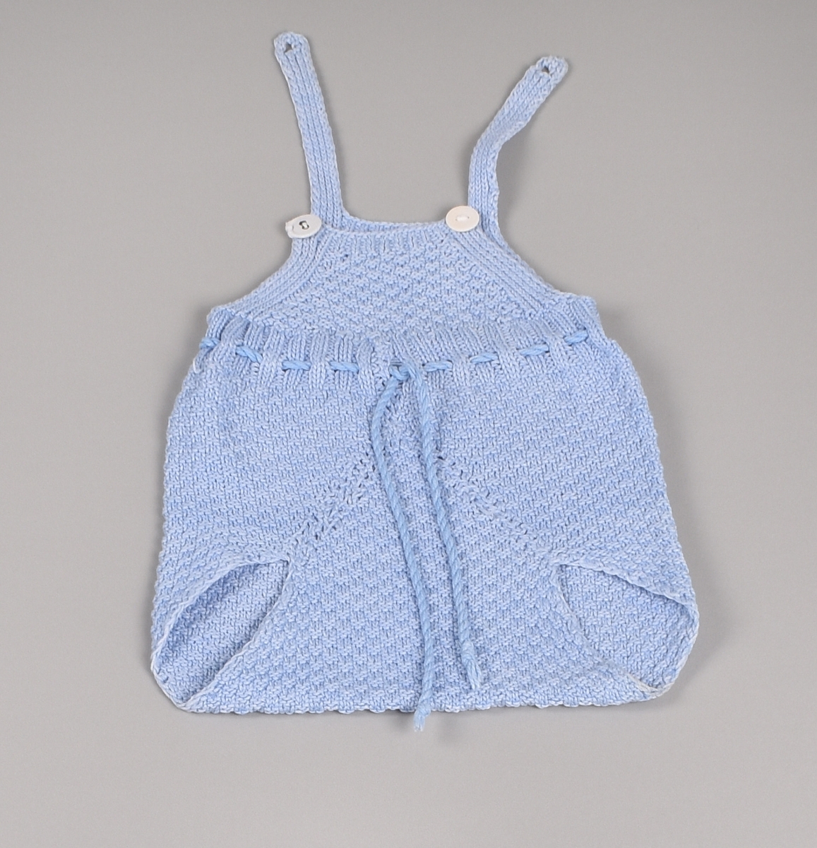 Lys blå strikket spedbarnsbukse med bærestykke og seler. Buksa er uten bein, har tvunnet snor i livet og selene festes foran med to knapper.