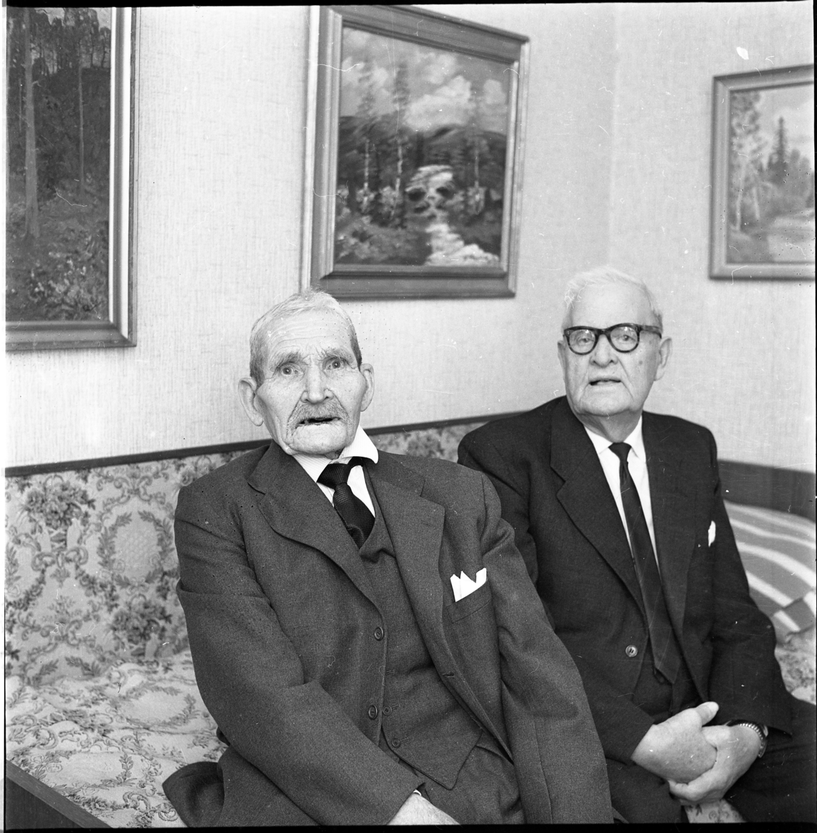 Två män sitter i en mönstrad soffa. Tavlor med skogsmotiv på väggen bakom dem. Mannen till vänster är 90-årige Karl Lindstrand i Lommaryd, troligen fotograferad i samband med hans födelsedag. Den något yngre mannen till höger är oidentifierad