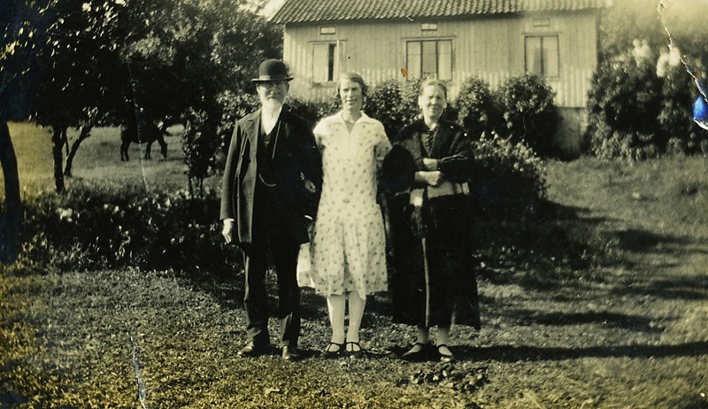 Familjen Magnusson poserar i sin trädgård vid "Fredriks" Hallen 1:2 (nuvarande Färåsvägen), okänt årtal. 
Från vänster: bonden Fredrik Magnusson (1855 - 1936), dottern Valborg "Bolla" Magnusson (1898 - 1970) samt hustrun Maria Magnusson (1863 - 1945). Boningshuset syns i bakgrunden. 
Relaterat motiv: A1662.