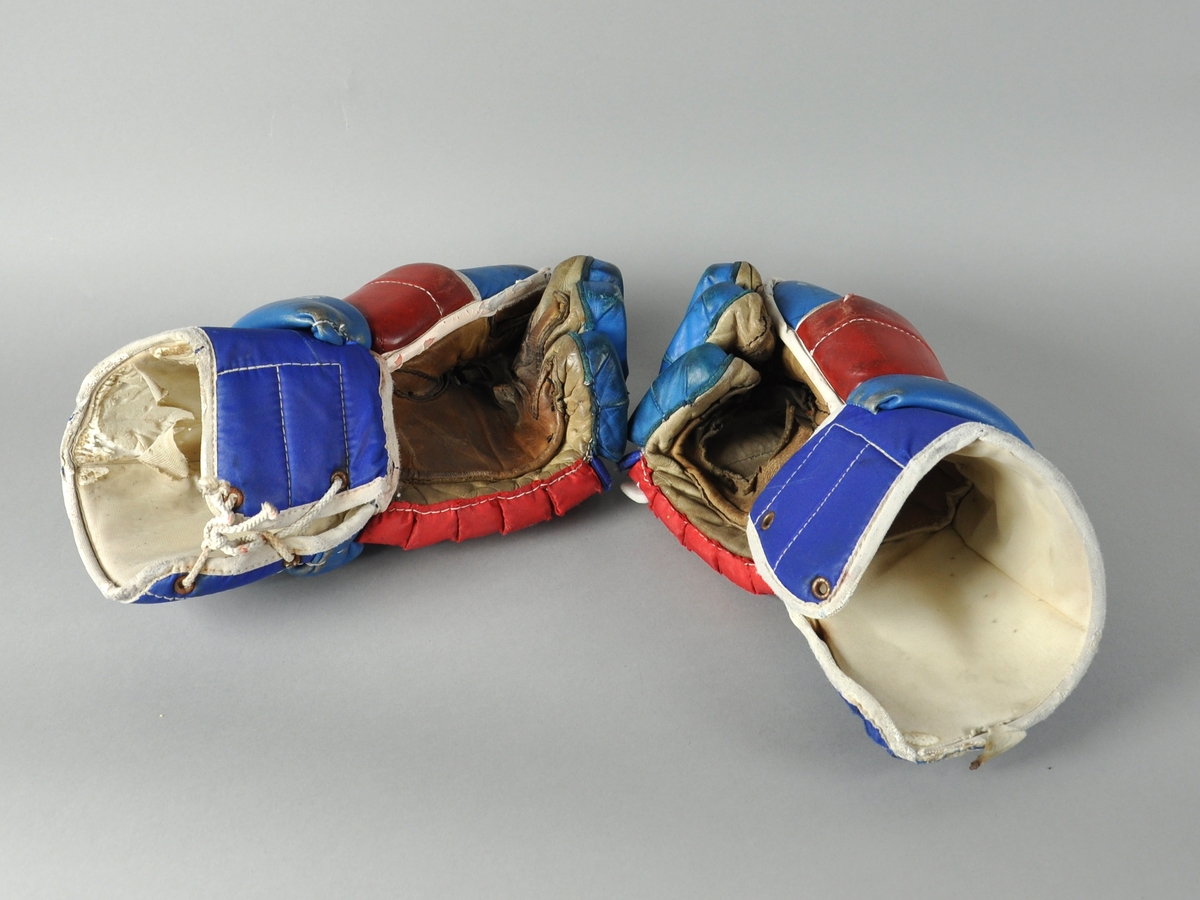 Et par Pallas ishockeyhansker av skinn i blått, rødt og hvitt. Hanskene er velbrukte og har flere hull på innsiden av hånden.