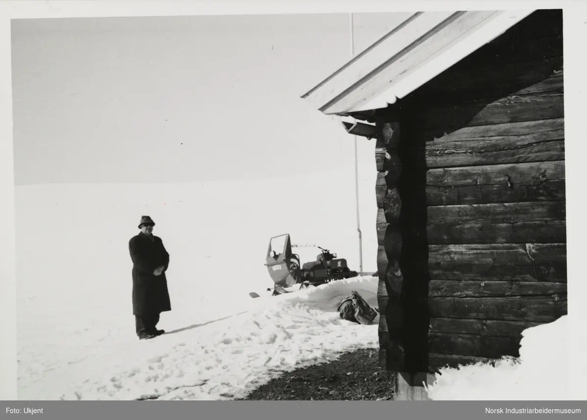 James Coward står i snøen ved snøskuter og hytte