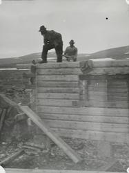 To menn står på laftet hytte under oppføring i fjellet