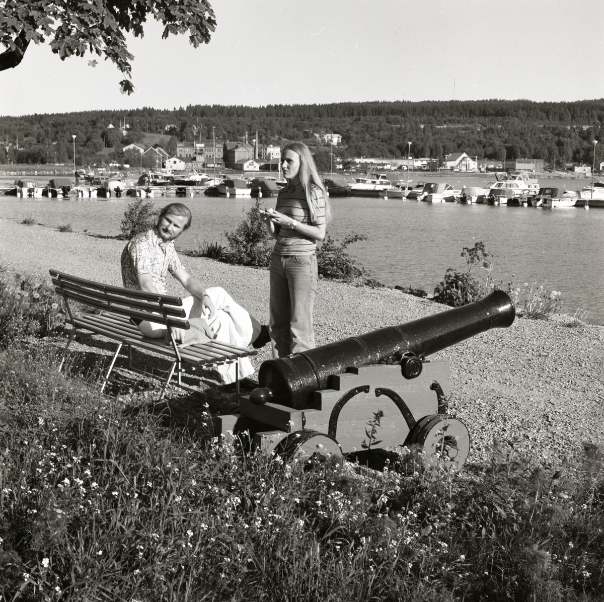 En man sitter på en bänk och en kvinna står bredvid. Intill henne står en gammal kanon och i bakgrunden syns en småbåtshamn.