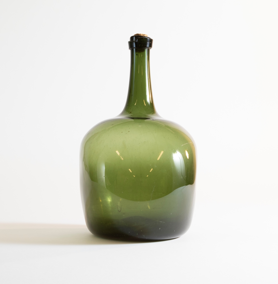 Flaska av grönt glas med tillplattad sfärisk form och hög hals. Del av kork sitter kvar i mynningen.