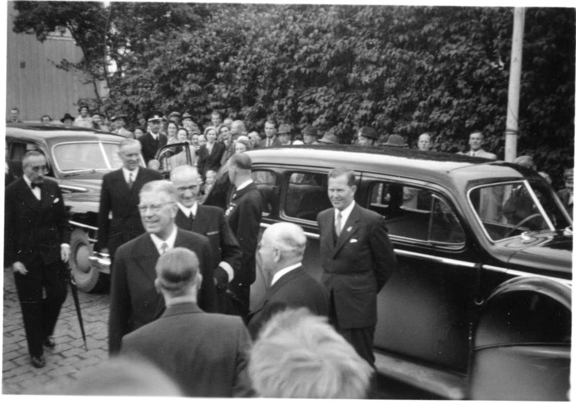 Kung Gustav VI Adolf med manligt sällskap på Brahegatan i Gränna framför stora svarta bilar. Bakom dessa, på trottoaren, står en folksamling.
