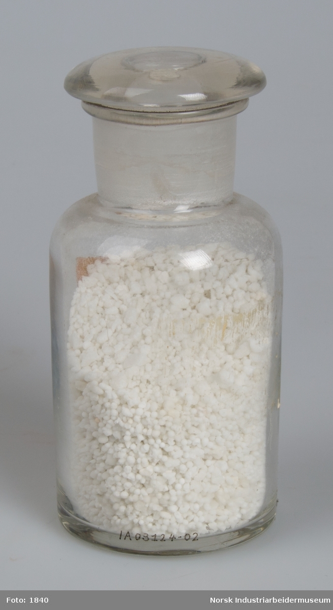 Laboratorieglas med prøve af den første produktion af kalksalpeter.
Cylindrisk laboratorieglas med slebet glasprop. Indeholder salpeter i form af små hvide perler og gryn.
