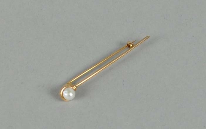 Stavbrosje av gull, med påmontert perle. Ligger i originaleske av papp. I gullstempelet er det motiv av det som ser ut til å være et vikingskip.
