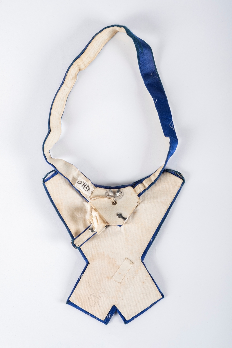 Sløyfe i blått tekstil med hvite geometriske motiver. Sløyfens ender ligger i kryss over hverandre. Under tekstilet er det papp som stiver av sløyfen, og en metallspenne der båndet rundt halsen kan festes.