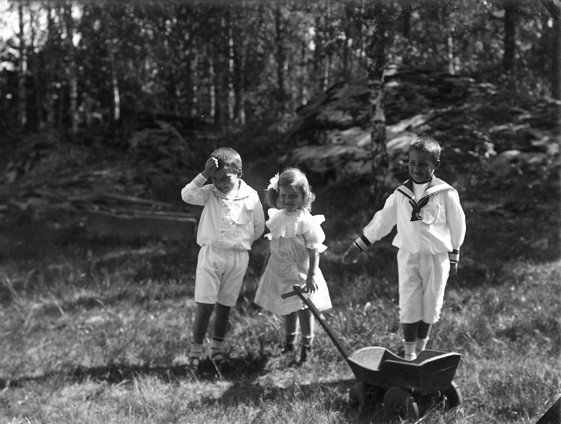 Barnen Stolpe från Stockholm. En liten flicka i ljus klänning står flankerad av två pojkar i sjömansuniform. Flickan håller en leksakskärra i handen.