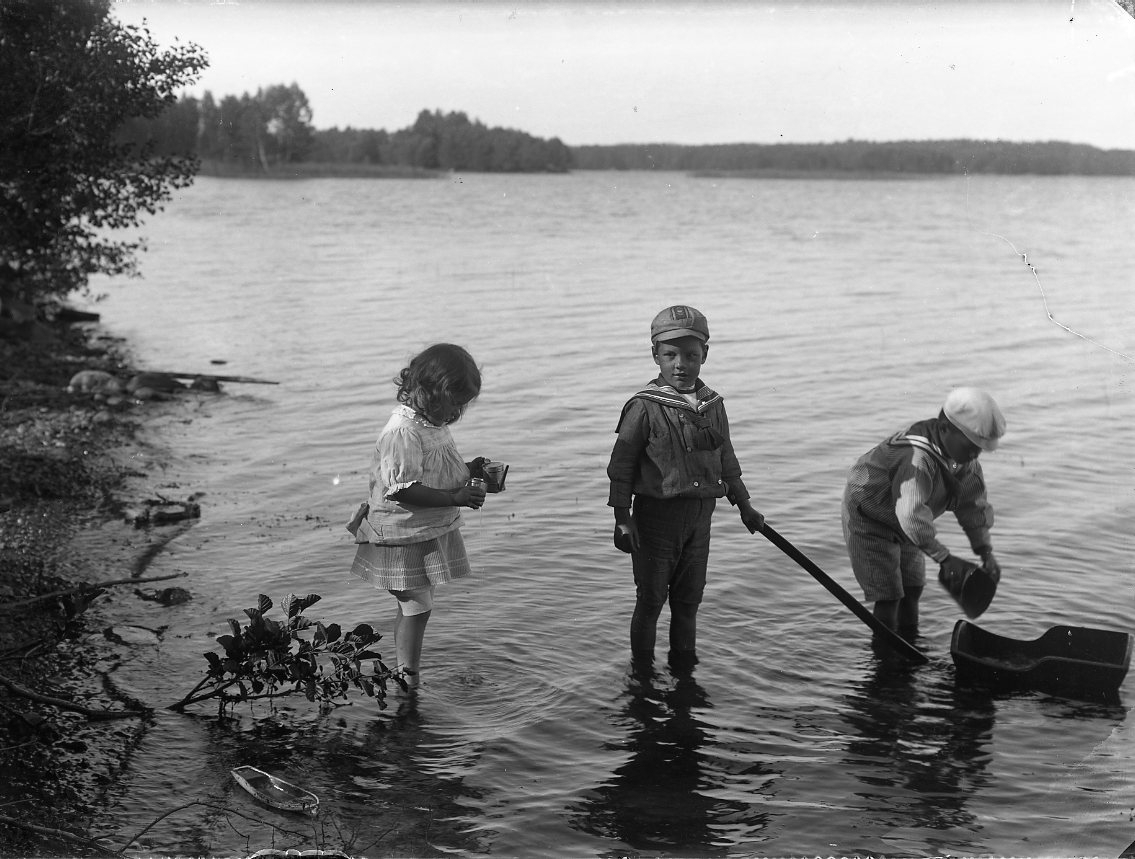 Barnen Stolpe från Stockholm står i vattnet vid en strand. En liten flicka i ljus klänning, något uppvikt, intill två pojkar i sjömansuniform som leker med att fylla en liten leksakskärra med vatten.