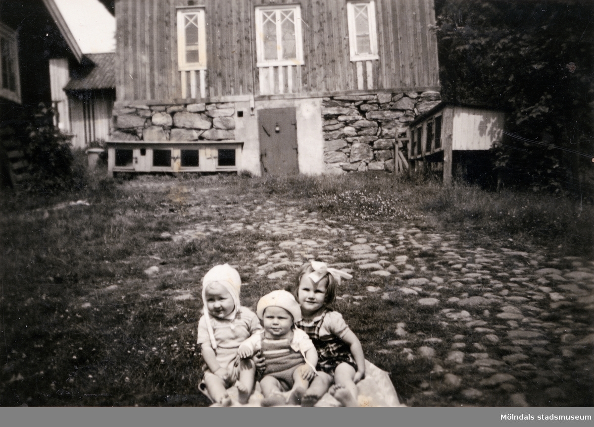 Porträtt av tre små barn på gården Christinedal i östra Mölndal på 1950-talet. I bakgrunden ses en av gårdens ekonomibyggnader. Gården har gett namn åt området Kristinedal och var belägen i vinkeln mellan nuvarande Rådavägen och Brovaktaregatan. År 1960 såldes gården till Fastighetsbolaget Betongblandaren (Skånska Cementgjuteriet) och revs när det nuvarande bostadsområdet anlades.