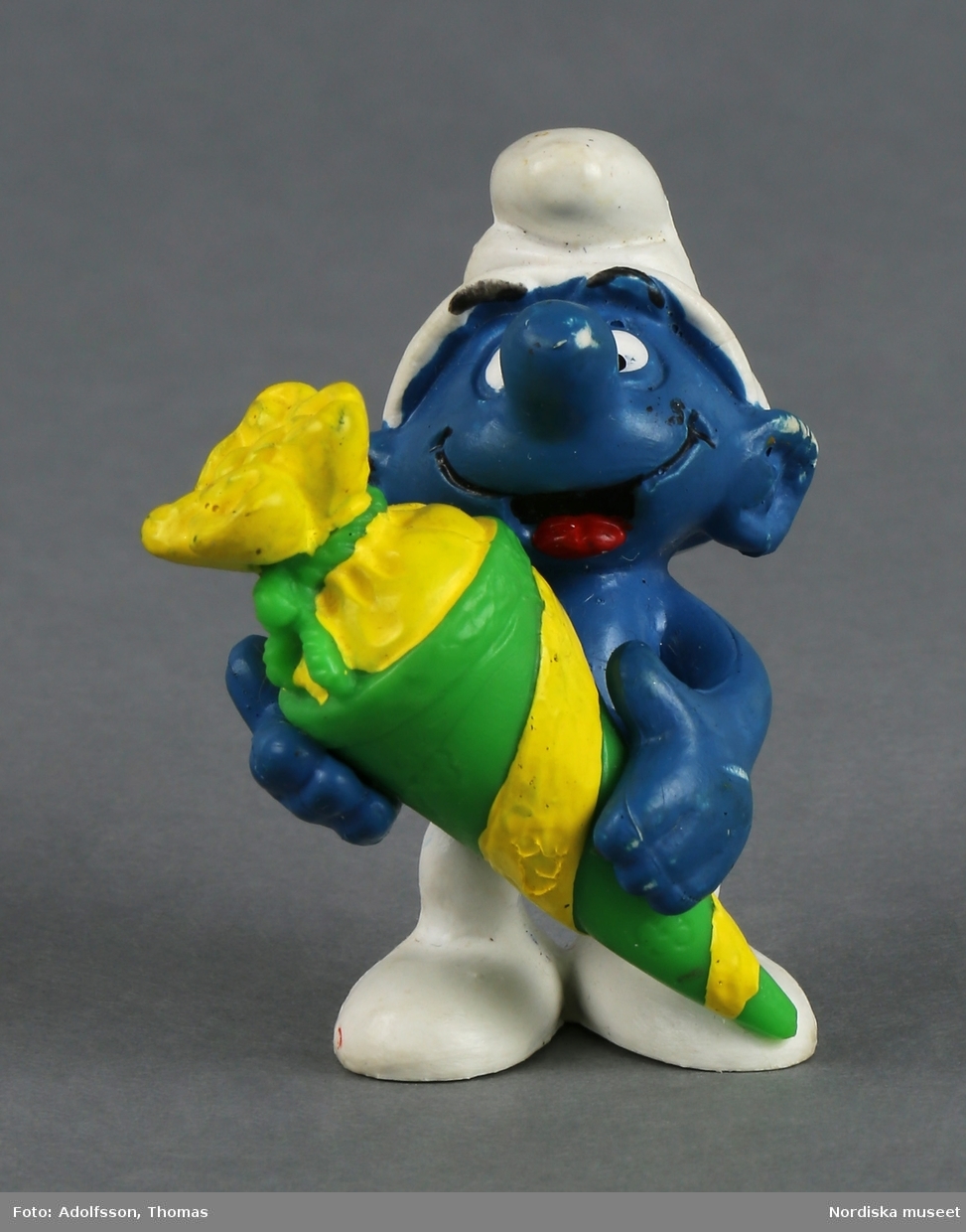 Tomteliknande figur som kallas Smurf, med blå kropp, vit luva och vita byxor. I handen håller han en stor gulgrön godisstrut.  Ansiktsuttrycket är glatt och tungan sticker ut.