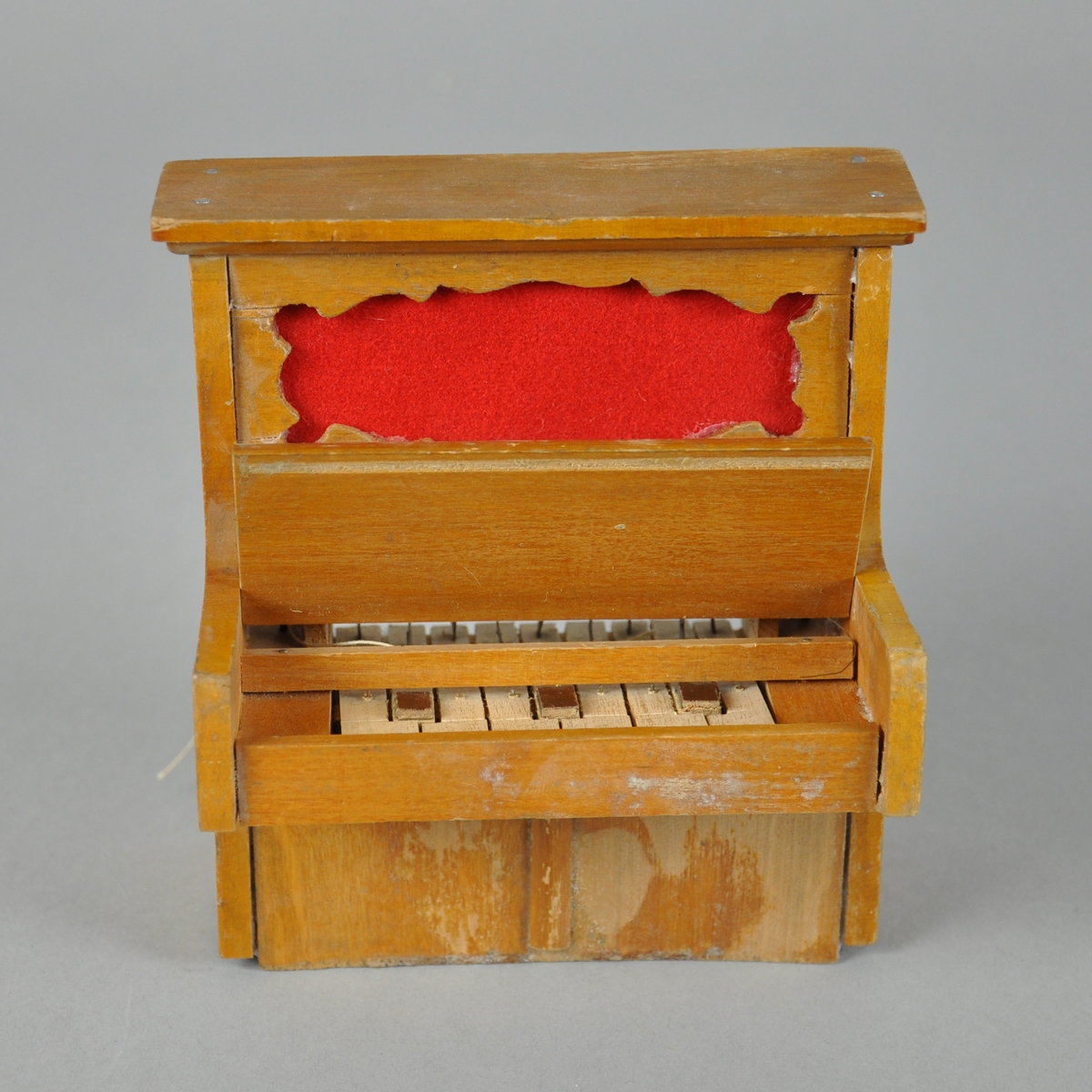 Lekepiano av tre, med utskåret overdel og rød filt. Pianoet har ni tangenter.