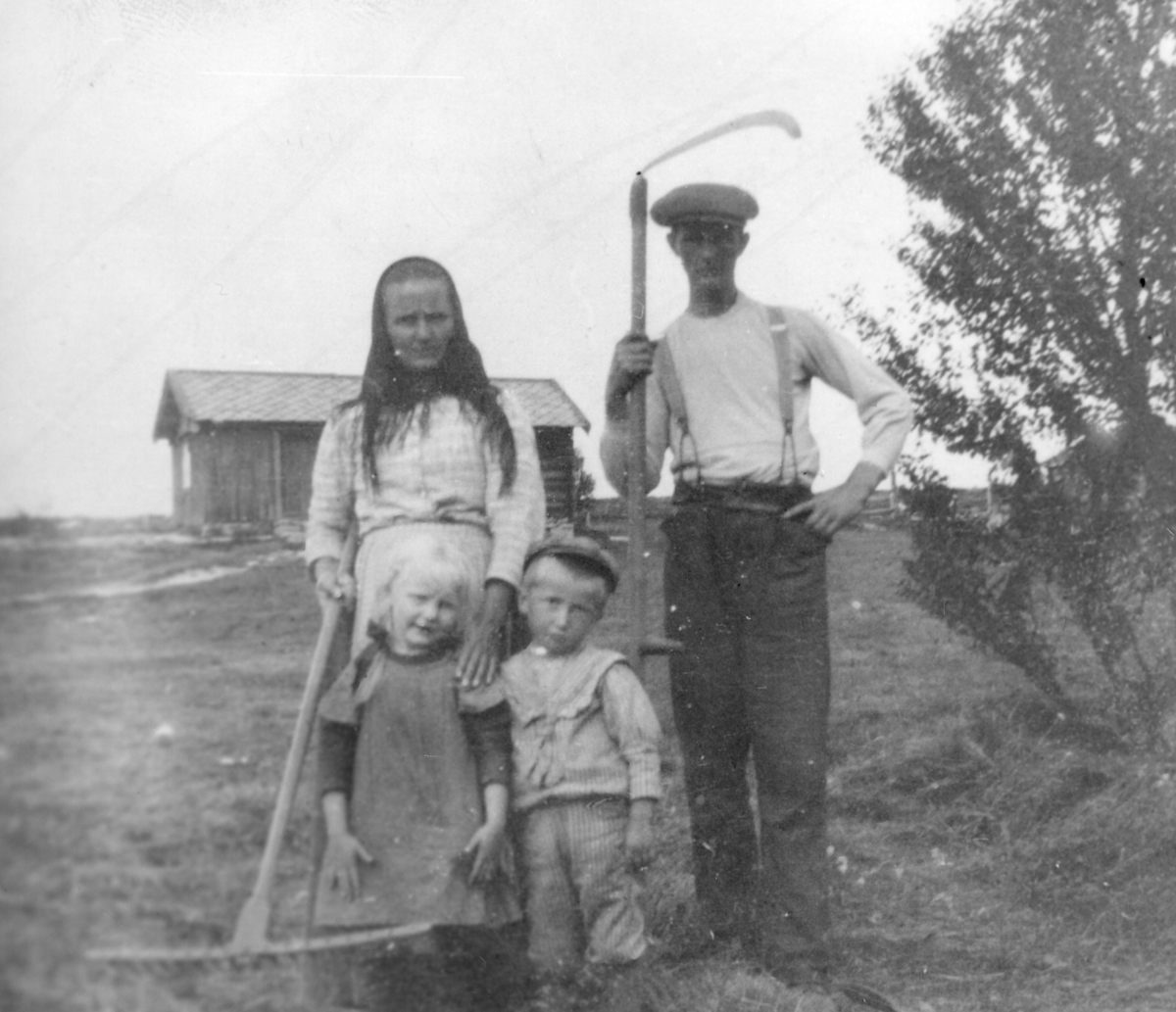 Vinne på Tronsbakken i Småsetran. Kvinne med rive og mann med ljå, samt to små barn poserer for fotografen