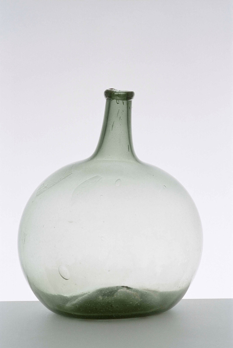 Flaska av handblåst grönt glas, bukig med tillplattade sidor. Hals med pålagd ring kring mynningen, svagt inbuktad i botten.
