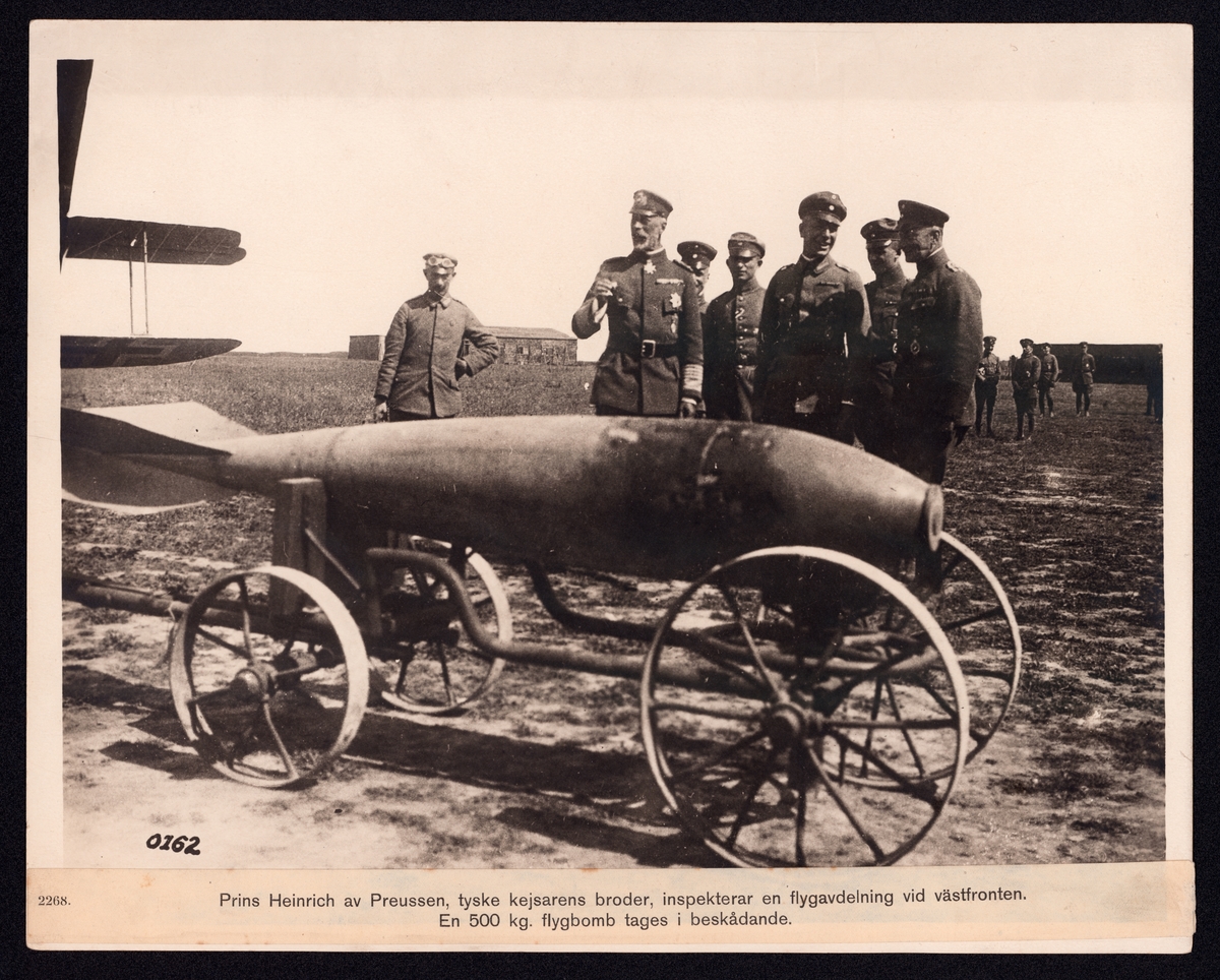 Bilden visar hur Prins Henrik av Preussen inspekterar en flygbomb på en transportvagn. Han är omgiven av en grupp officerare och ett flertal soldater som håller sig i bakgrunden. På vänster sidan syns vingarna av ett dubbeldäckarflygplan.
