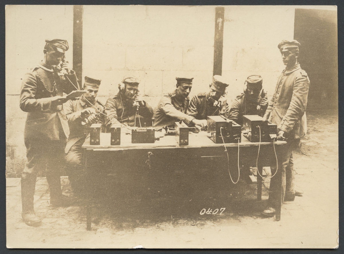 På bilden syns en grupp soldater som sitter runt ett bord med telefonapparater och håller på att föra telefonsamtal. Mannen till höger verka vara en officerare som håller uppsikt. På vänster sidan finns också en officerare som har tagit över ett samtal.