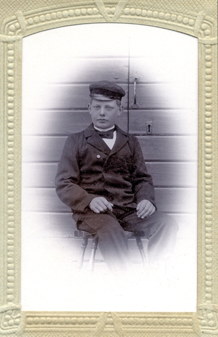 Foto av en ung man (tonåring) klädd i jacka, skärmmössa och fluga. Han sitter på en pinnstol framför en ytterdörr. Bakpå fotot står: "Gustaf Lönn 14 år gammal". 
Knäbild, en face.