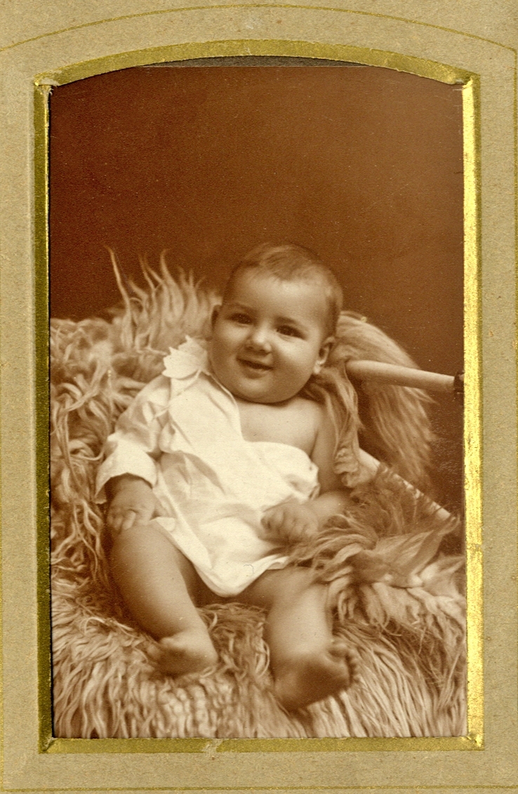 Foto av ett spädbarn i kolt, som sitter på en fäll. 
Helfigur. Ateljéfoto.