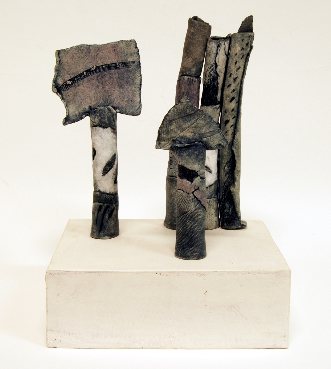 Skulptur i stengods på sockel av kaolinbestruken terrakotta. Skulpturen består av tre skilda vertikala "pelare" avslutade upptill (två av dem) med en tvärställd skiva. Mönstrad gråblå yta. Signerad nedtill på podiets kortsida: "Ninna Gunnarsson".