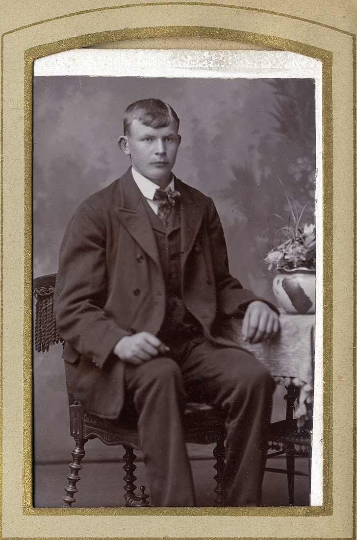 En ung man i kostym med väst, som sitter på en stol vid ett bord med en blomkruka på. 
Knäbild, halvprofil. Ateljéfoto.