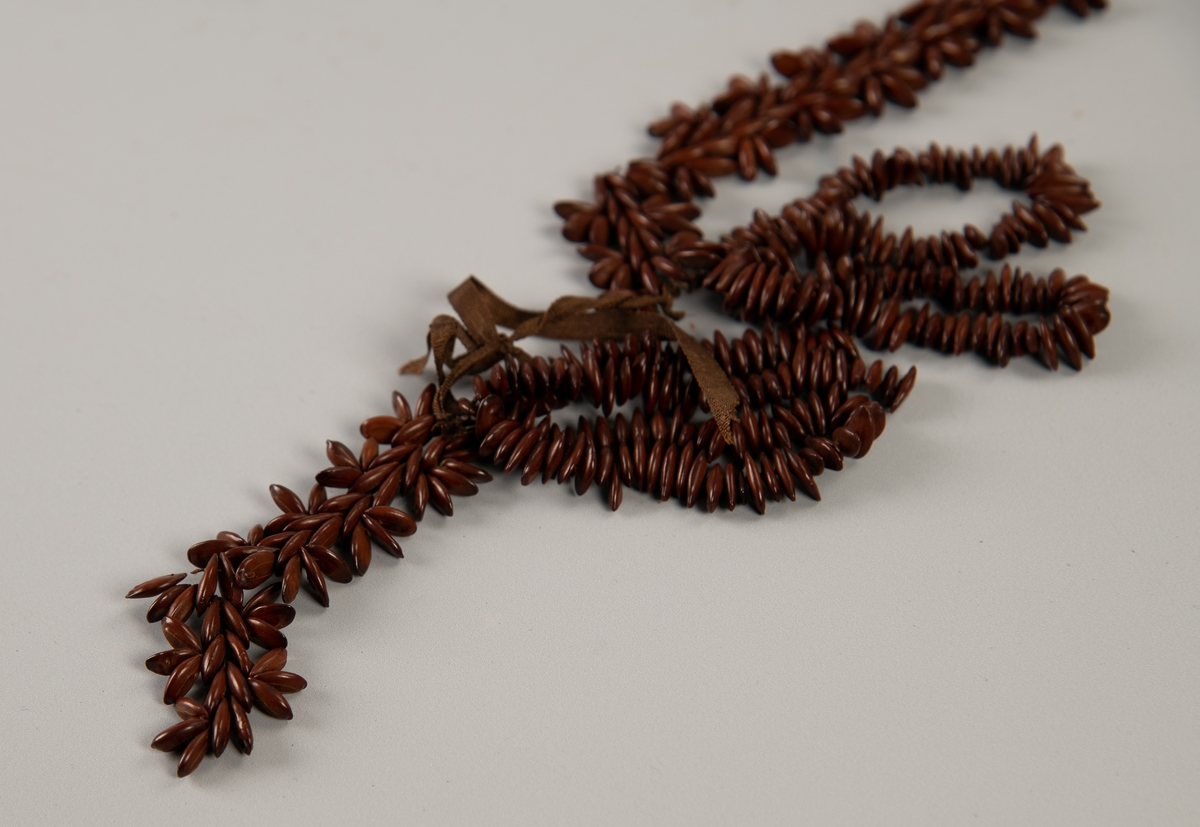 Armbånd  fra St. Helena. Sjokoladebrune   fruktkjerner, tynt, brunt  silkebånd. Ca. en halv cm. lange ellipseformede kjerner tredd på tynt, brunt silkebånd.