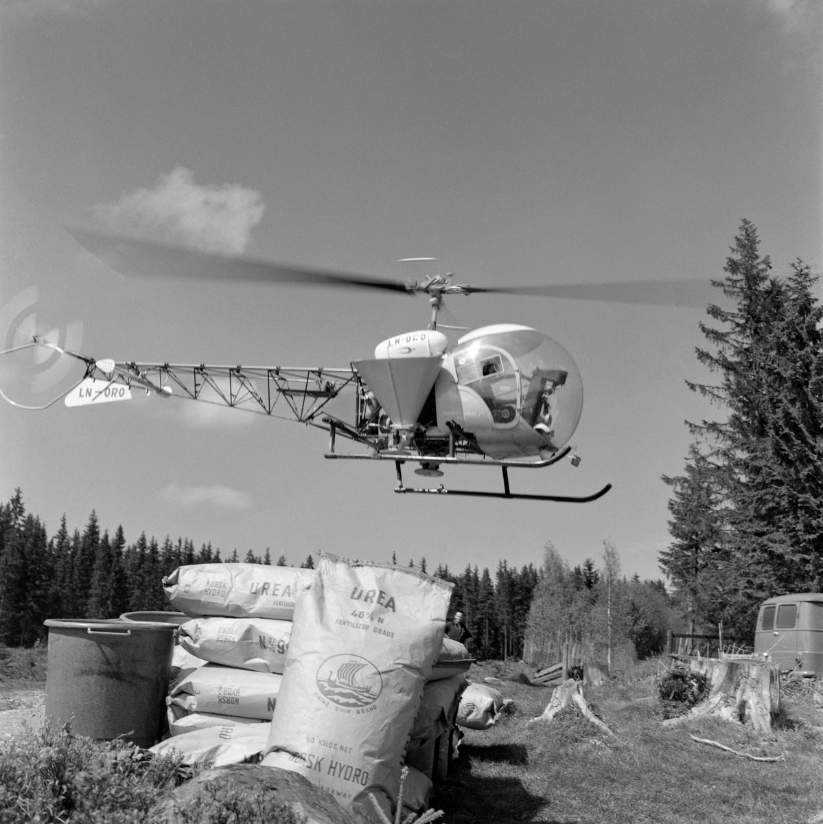 Forberedelser til gjødsling av skog fra helikopter av typen Bell 47. Fotografiet viser et helikopter med sidemontert gjødselbeholder i lav høyde over bakken over ei åpen flate i skogen. I forgrunnen ser vi en del sekker med «Urea» - ei gjødselblanding som ble utviklet spesielt for det norske skogbruket av Norsk Hydro, som et ledd i bestrebelsene for å øke tilveksten i de norske skogene med sikte på å være mest mulig sjølforsynt med råstoff til trelast- og papirindustrien. Gjødslingsaktiviteten ble orientert mot ungskogen, for det gjaldt å sikre den nevnte industrien noenlunde jevn råstofftilgang.  Skogeiersamvirket bidrog med å skaffe i traktormonterte spredeaggregater. Tidlig i 1960-åra etablerte Felleskjøpet en avdeling som tilbød gjødsling av skog fra fly.  Selskapet tok oppdrag så fremt det var snakk om sammenhengende areal på minst 100 dekar og start- og landingsmuligheter for småfly på en rett vegstrekning eller en innsjø mindre enn tre kilometer fra bestandet. Da dette fotografiet ble tatt var tydeligvis også helikopterspredning blitt et aktuelt alternativ. Landbruksdepartementet åpnet muligheter for finansiering av gjødslinga gjennom skogavgiftsordningen. Landbruksdepartementet åpnet muligheter for finansiering av gjødslinga gjennom skogavgiftsordningen. Med et slikt apparat på plass ble 90 000 dekar norsk skogsmark gjødslet i 1967. Det var primært store skogeiendommer som ble gjødslet, og forkjemperne for denne virksomheten så på eiendomsstrukturen i norsk skogbruk, med mange små teiger, som en begrensende faktor. Det viste seg imidlertid etter hvert at prognosene for den volumtilveksten skogen skulle få etter gjødsling hadde vært vel optimistiske. Derfor avtok denne aktiviteten utover i 1970-åra. Skoggjødsling fikk en renessanse i 2016. Da ble det kjent at staten gav tilskudd som dekte 40 prosent av gjødslingskostnadene i ensaldret, eldre barskog, bestand som skulle avvirkes i et tiårsperspektiv. Bakgrunnen for denne skogpolitiske kursendringa var ikke bare en vilje til å gi næringa økonomiske stimulanser – det handlet også om at skog i god vekst er en viktig faktor i arbeidet med å begrense de klimaproblemene som skapes ved store utslipp av karbondioksid i atmosfæren som følge av utstakt bruk av fossile energikilder.