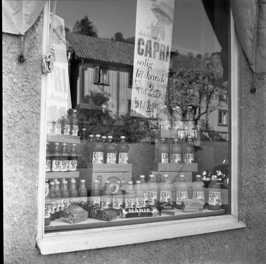 Fotografi av skyltfönster till Firma Gustaf Rahm HB på Brahegatan 61. I fönstret står det flaskor med Capri Apelsin - troligen saft - och en reklamskylt med texten: "Drick Capri solig läskande 1/1 2:15 1/2 1:20". I fönstret speglar sig vandrarhemmet mittemot, Brahegatan 56.