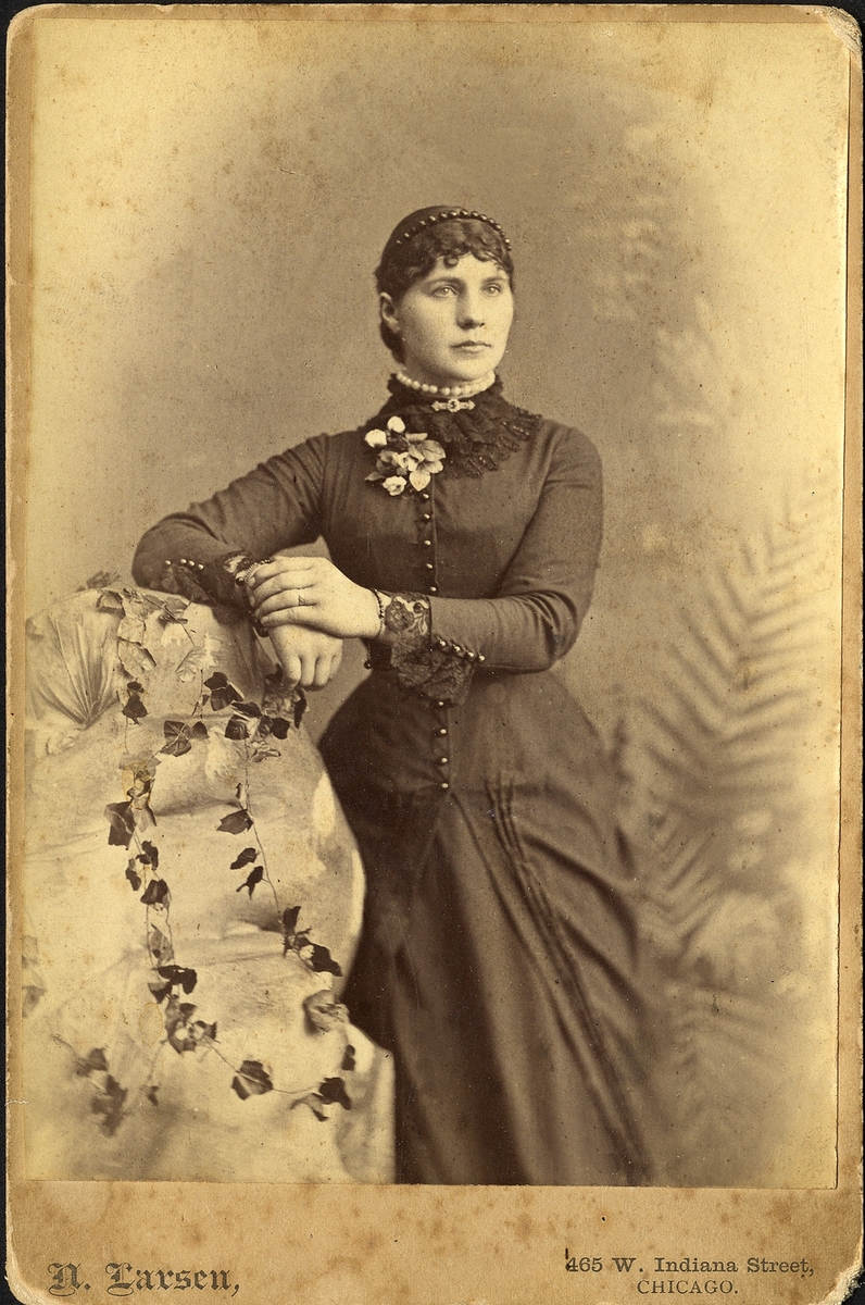 En okänd kvinna i mörk klänning med antydan till turnyr. Vid kragen syns en brosch och ett tättsittande halsband. I håret sitter ett stenkolsdiadem. 
Knäbild, halvprofil. Ateljéfoto.