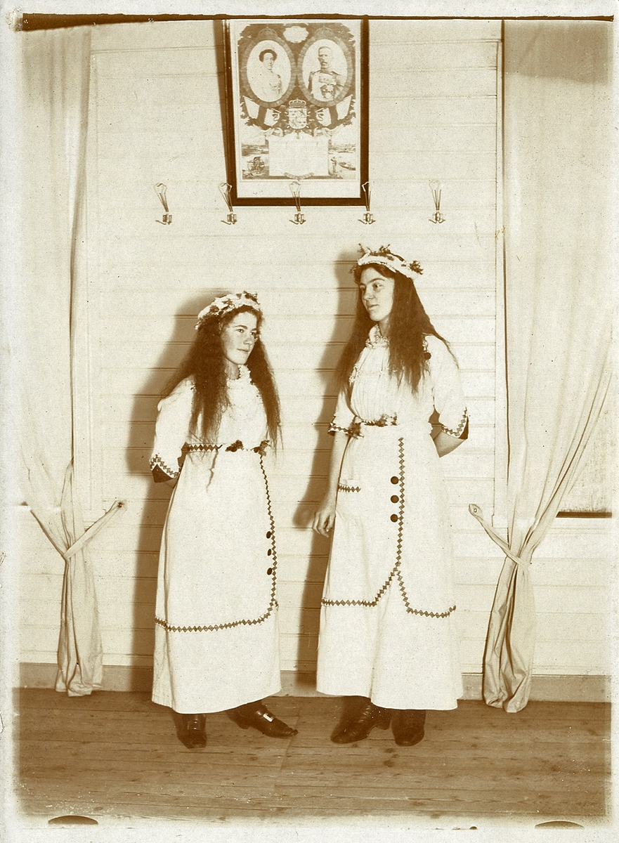 Två utklädda tonårsflickor. På väggen bakom dem syns en tavla med Gustaf V och drottning Viktoria.