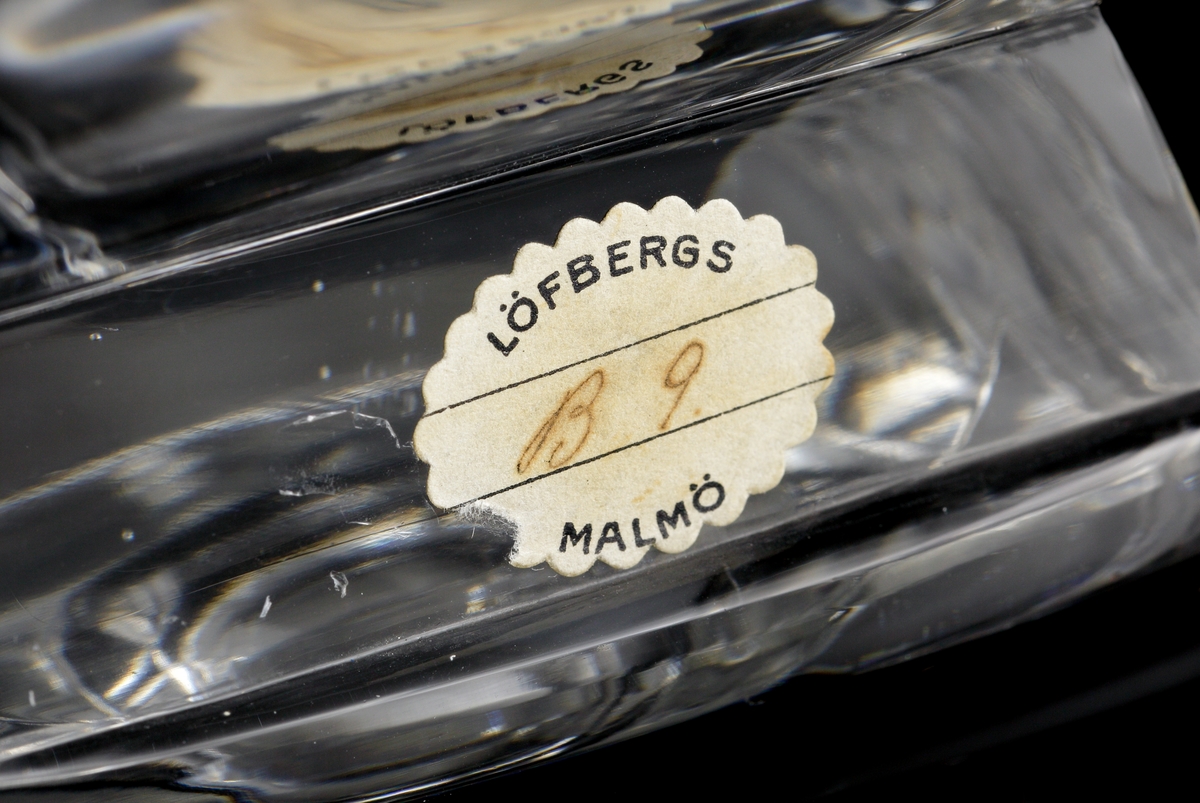 Vas med fyra sidor som slipats till sin fyrkantiga form. Ofärgat klarglas.
Etikett: Från Löfbergs Malmö. Handskrivet i mitten: "B 9."
