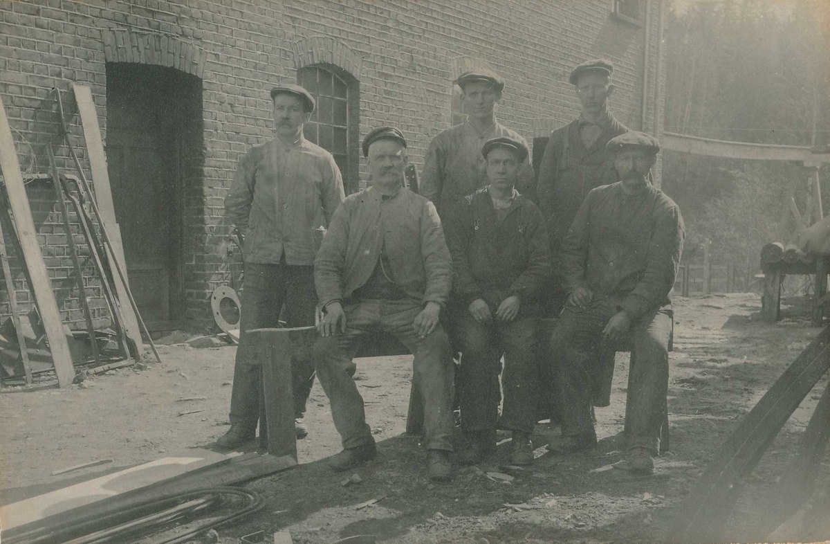 Gruppebilde av seks sittende og stående arbeidskledde arbeidere utenfor industribygning i tegl.