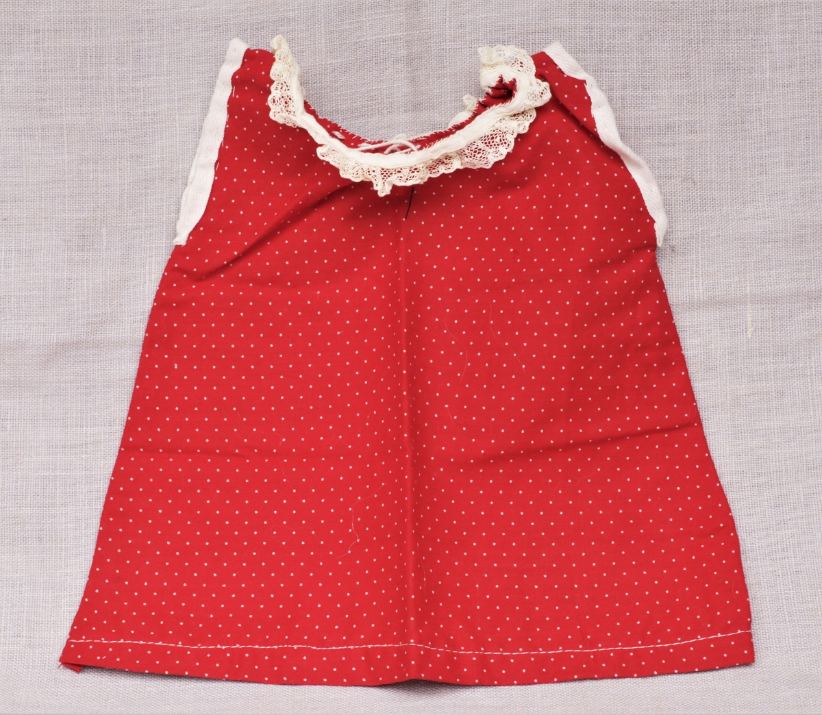 Tre enkle dukkekjoler, to rødprikkete (a og b) og en blårutet (c), en stråhatt (d), et bånd (e). Rød kjole (b) uten ermer, med hvite kanter.