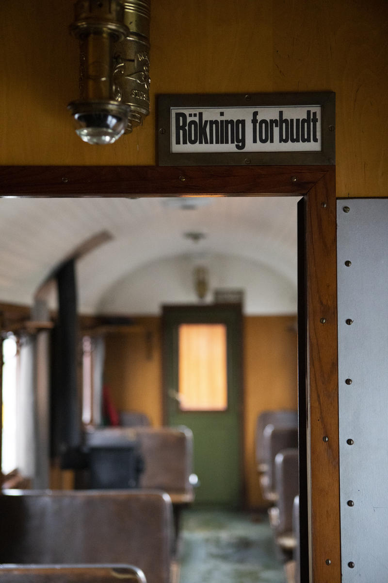 Interiør i en av Urskog-Hølandsbanens passasjervogner for tredje klasse. I forgrunnen ses et emaljeskilt med teksten Røkning forbudt. Det er sort tekst på hvit bakgrunn. I bakgrunnen ses vognens passasjerseter trukket i brunt skinnlignende stoff.