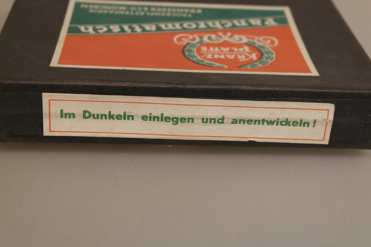 Tre esker med fotoplater. Eskene er fra tre ulike produsenter og er uåpnet. Alle inneholder plater i str 12 x 16,5 cm.
A) Svart eske fra Kranseder & Cie i München. Esken er pålimt en rød, grønn og hvit etikett på lokket, samt mindre etiketter på tre av sidene, alle med tysk tekst.
B) Lys brun eske fra Kodak. Esken har rød og svart tekst, og er pålimt en gul etikett med grønn tekst. Teksten er på engelsk.
C) Lys brun eske fra Ferrania. Esken har svart tekst på italiensk med grønn ramme rundt, og åpnes med en klaff på undersiden som er pålimt en rund etikett. Holdbarhetsdato oktober 1943.
