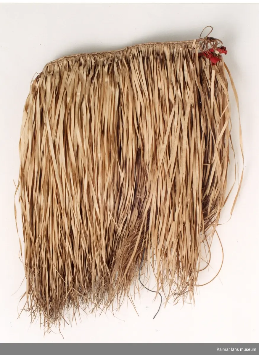 KLM 1950. Kjortel eller midjebeklädnad. Av gräs- eller vassblad, sammanflätade med kokosbast, prydd med röda tyglappar.