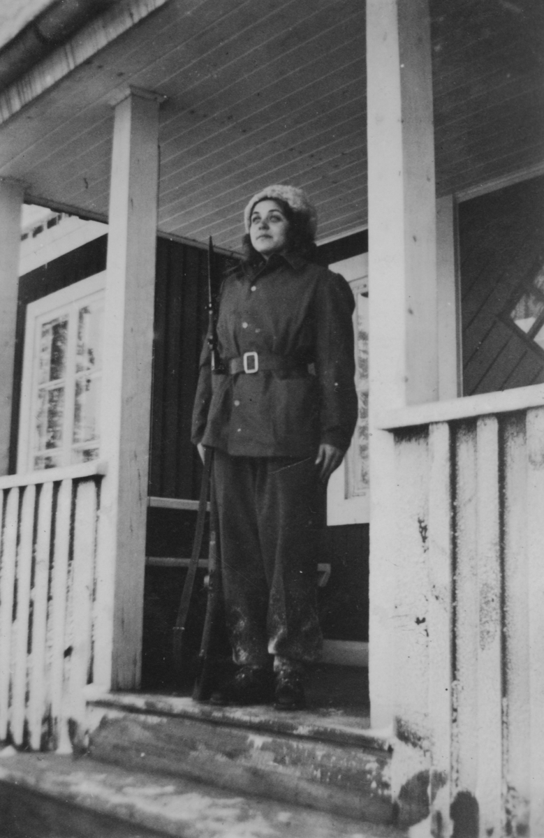 Porträtt av luftbevakare Inger (efternamn okänt) ståendes i givakt, iklädd uniform och med gevär, vintern 1942. Utomhus på verandan till luftbevakningsstationen för 91:a ls-kompaniet i Tellejåkk, Kåbdalis under beredskapsåren.