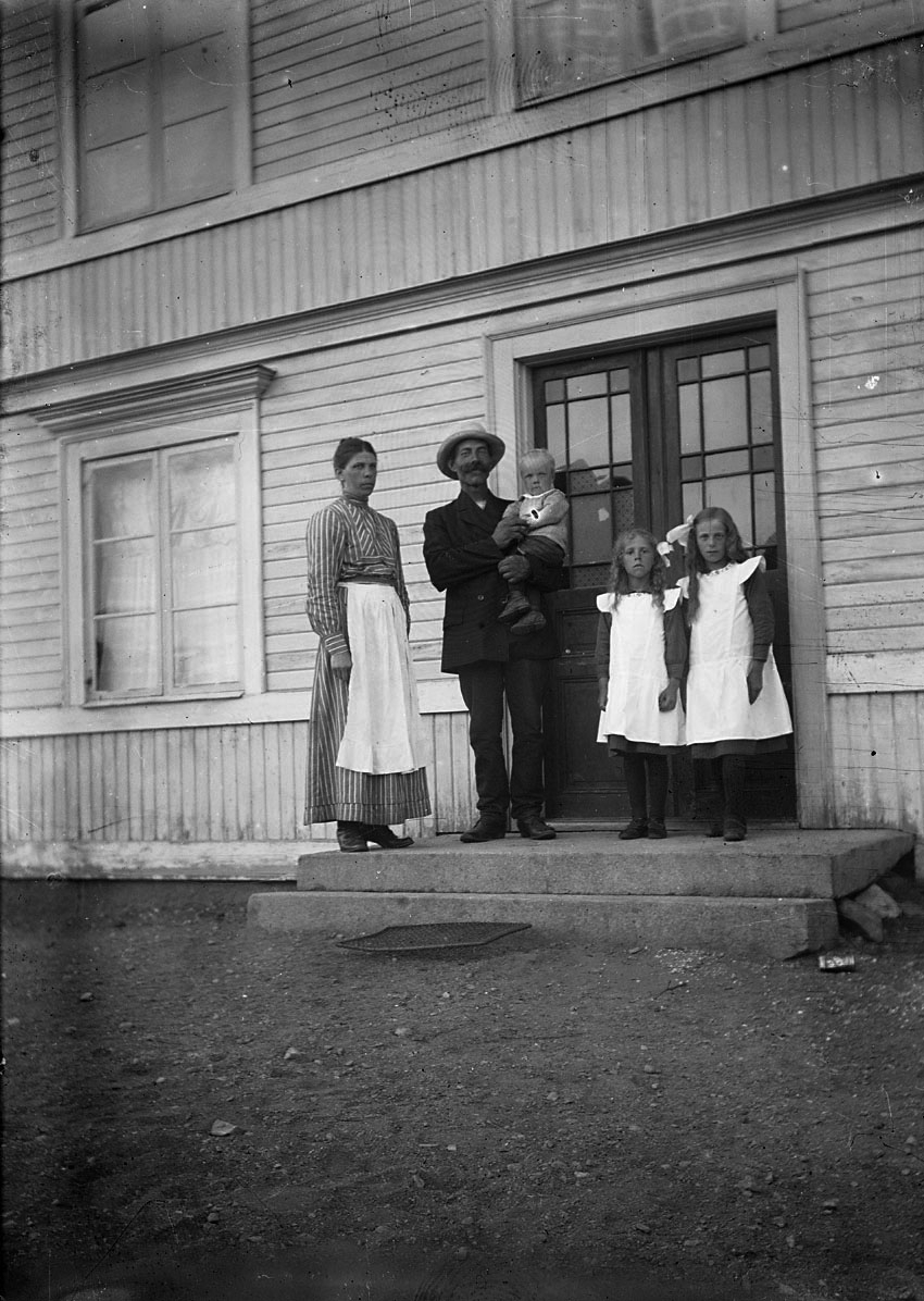 Par med tre barn, stående på trappa till hus.