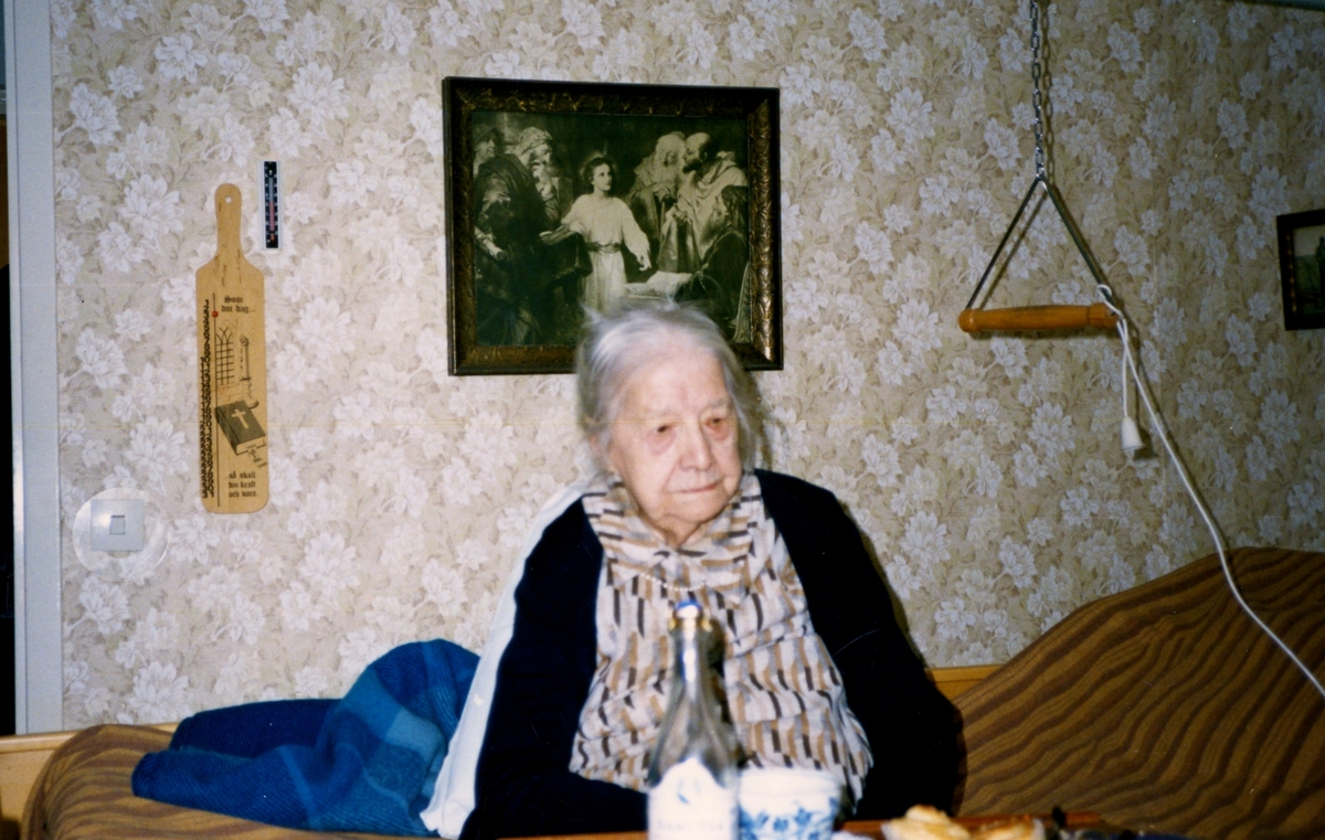 Ester Kristensson (1888 - 1990) sitter i en säng på Brogården, okänt årtal. Ester arbetade mycket inom kyrkan och i syföreningen. Hon var gift med Ivar Kristensson (1887 - 1976) som var kyrkvaktmästare/kyrkvärd.