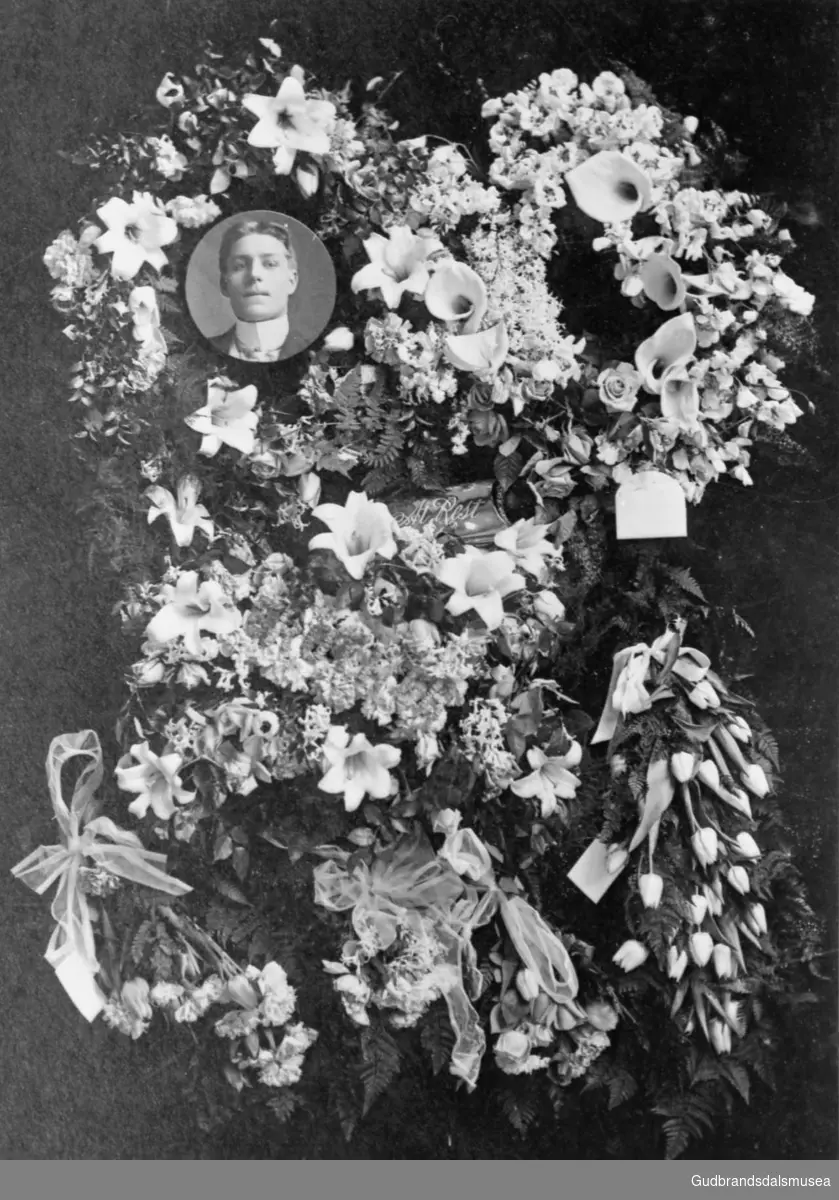 Lite portrettbilde av Syver Enebo er plassert i en blomsteroppsats, mange blomsterkranser rundt. Til minne om?