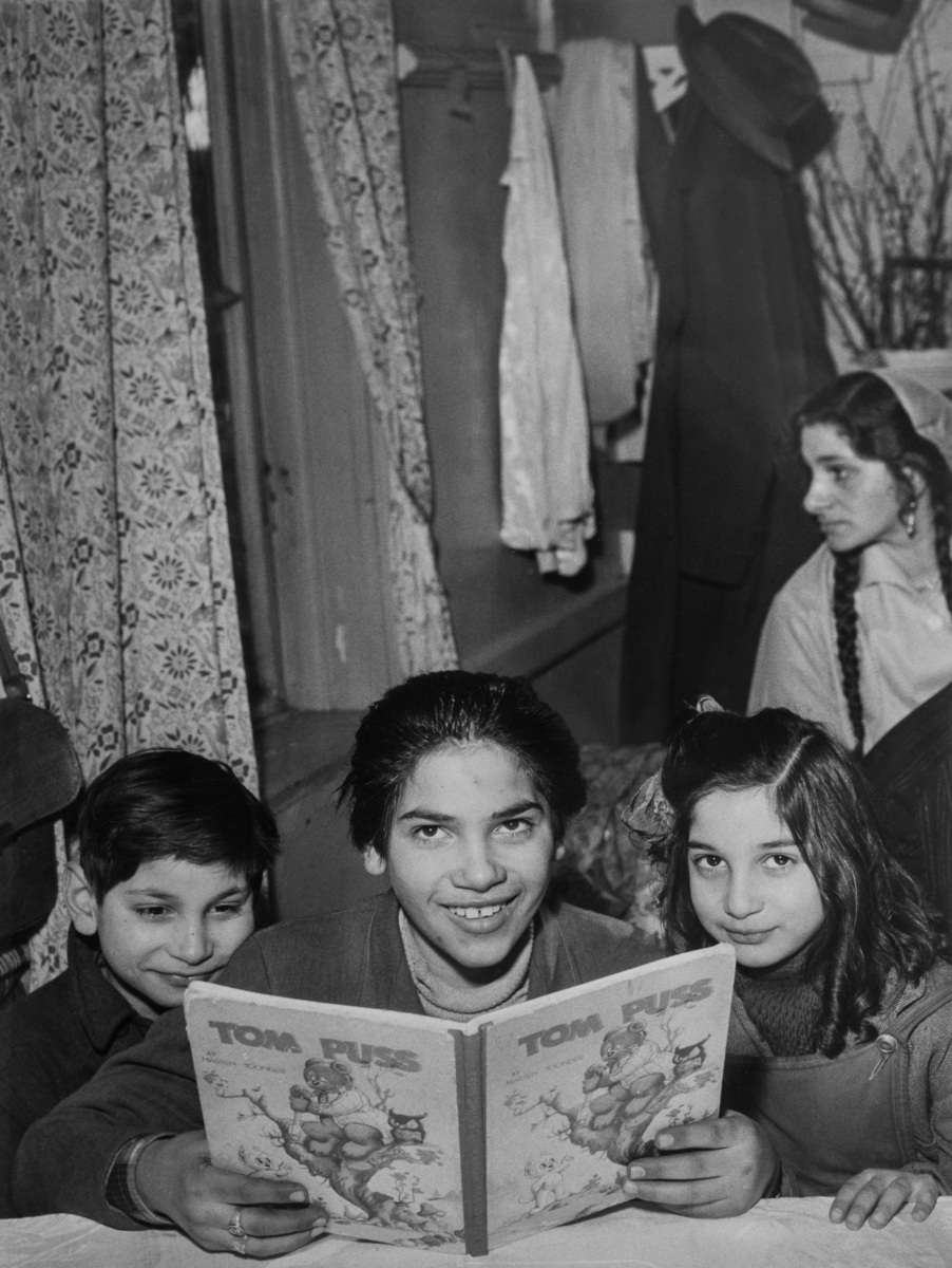 Tre barn poserar bakom en bok. Bilden är troligen tagen inuti en bostadsvagn.