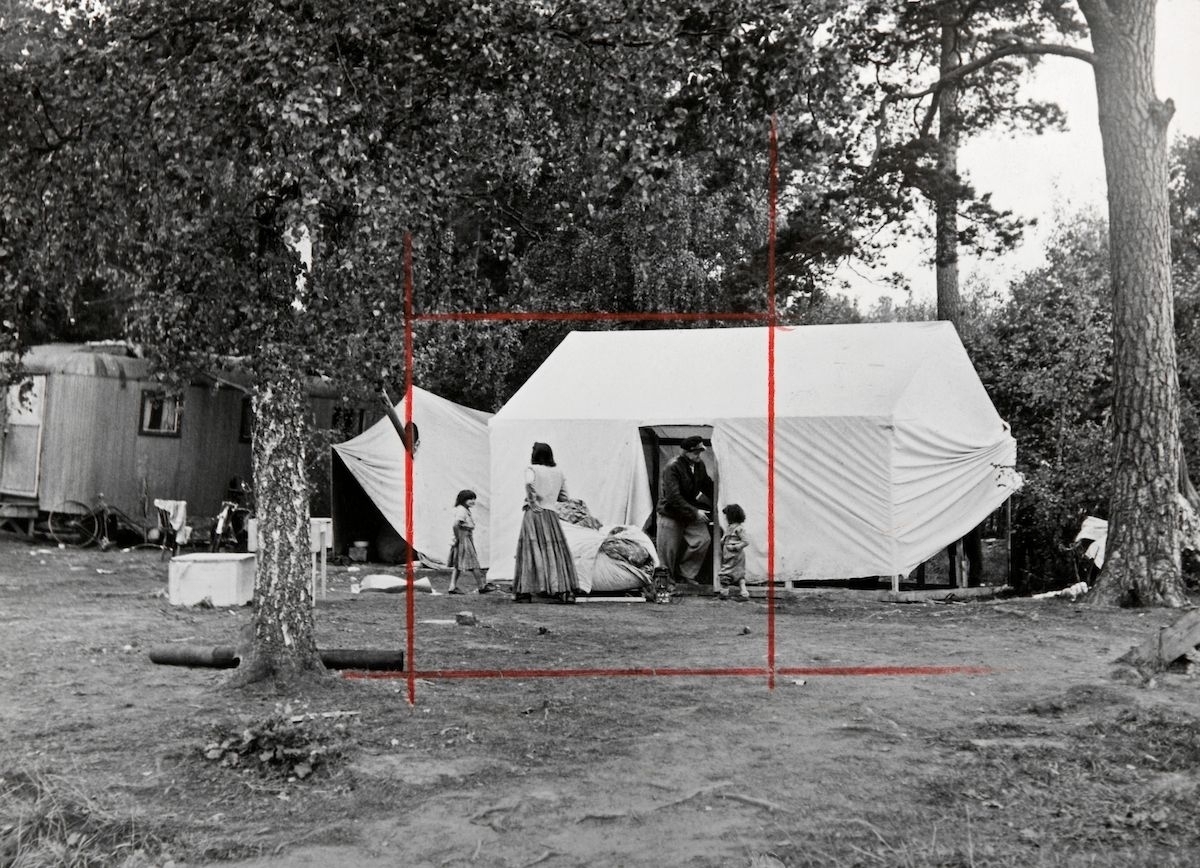 Bilden är troligen i tagen i samband med ebn vräkning av romer i Mälarhöjden, Stockhom, år 1951. Utanför ett tält står en man, en kvinna och två barn. Runt dem ligger täcken och saker på marken.