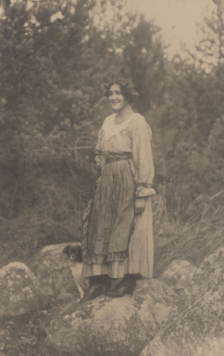 En kvinna står vid ett skogsparti med en hund vid sin sida.